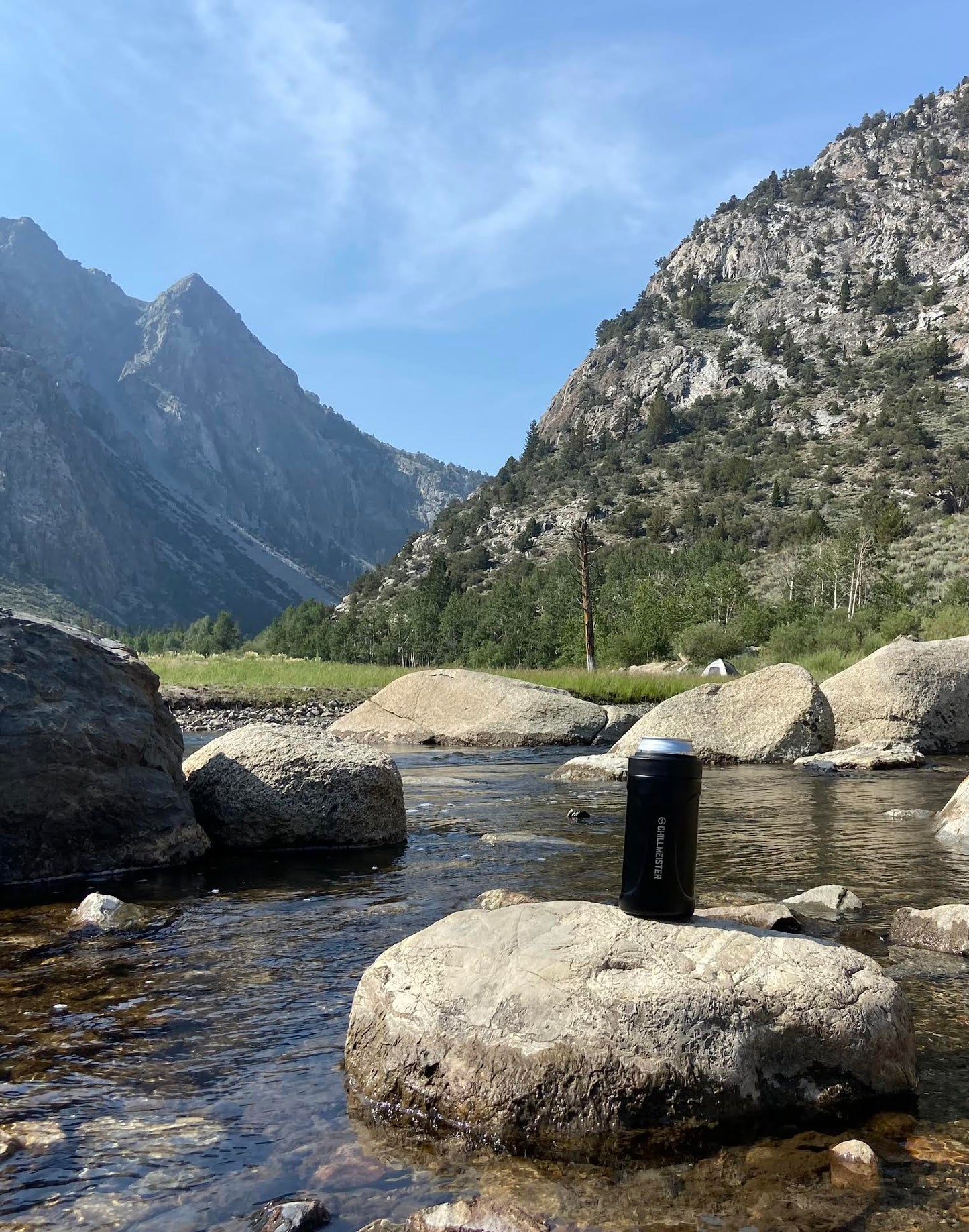 A mountain view scenery with a small river with large rocks. One rock has the matte black can cooler on top of it