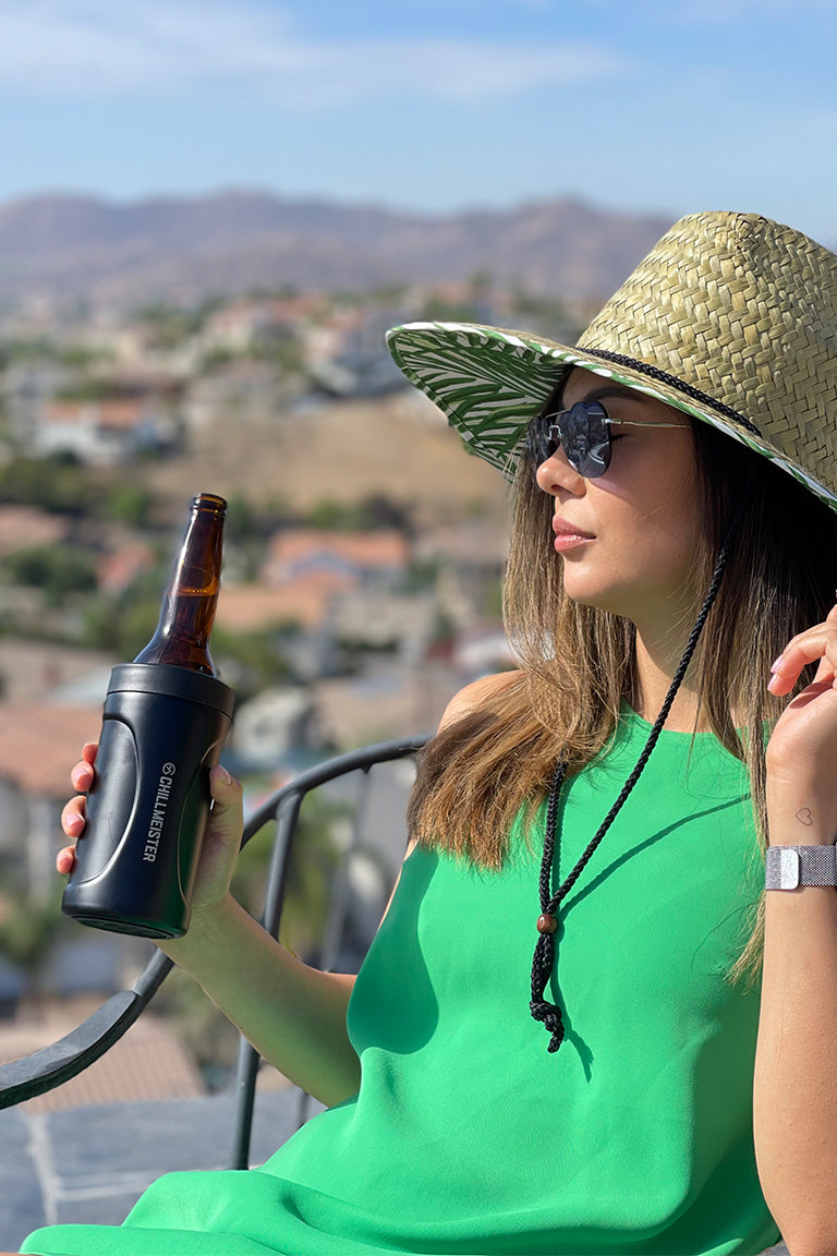A photo of a female model holding a black can cooler with a bottle inside about to take a sip while wearing a green dress and a straw hat