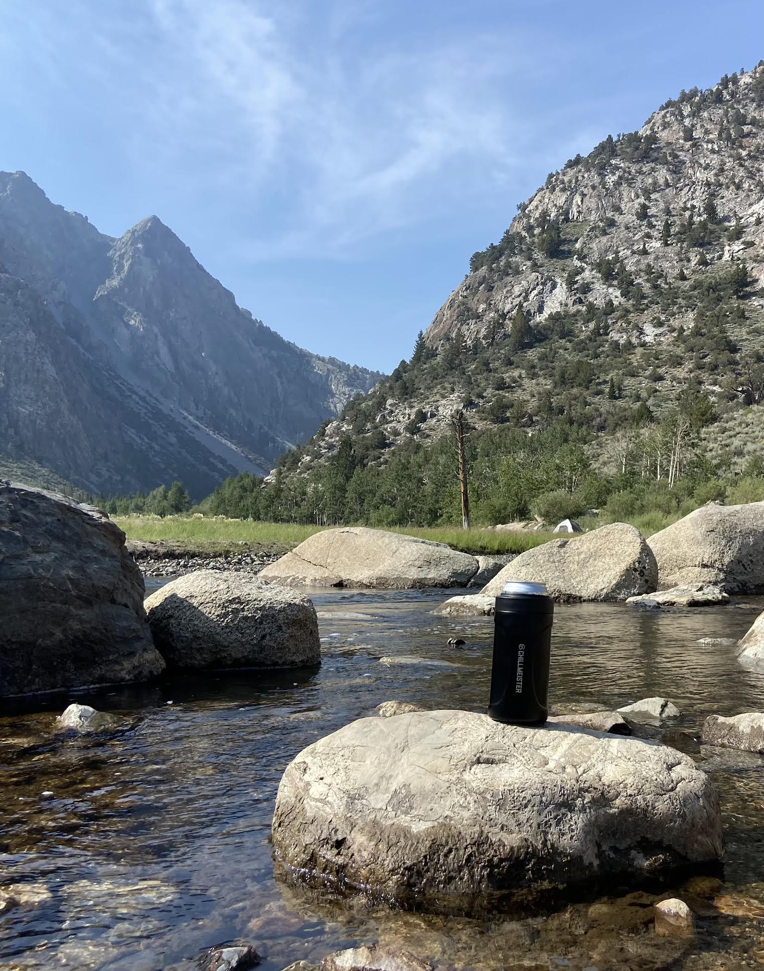 A mountain view scenery with a small river with large rocks. One rock has the matte black can cooler on top of it