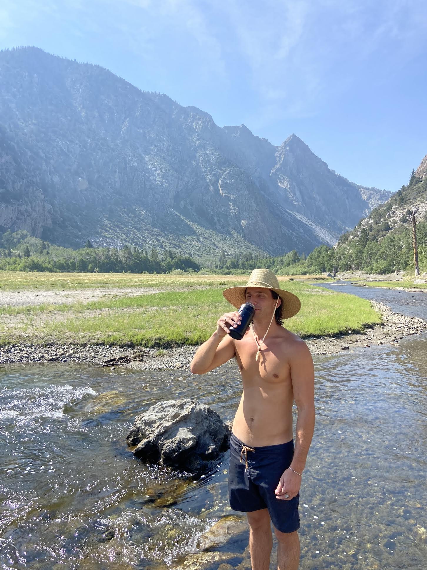 A male model wearing a straw hat while drinking a can cooler outside in front of a mountain view