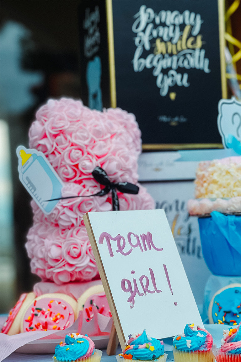 A close up shot of a pink foam covered flower bear with a sign it's a girl on a table with cupcakes and gifts