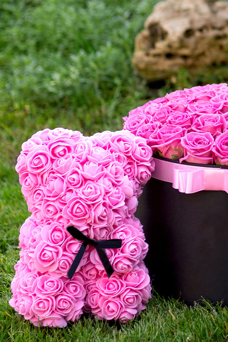 A close up shot of a pink foam covered flower bear next to a basket of pink roses on green grass