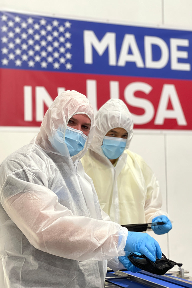Two people wearing protective clothing and blue face mask in front of a sign with the words "Made in USA"